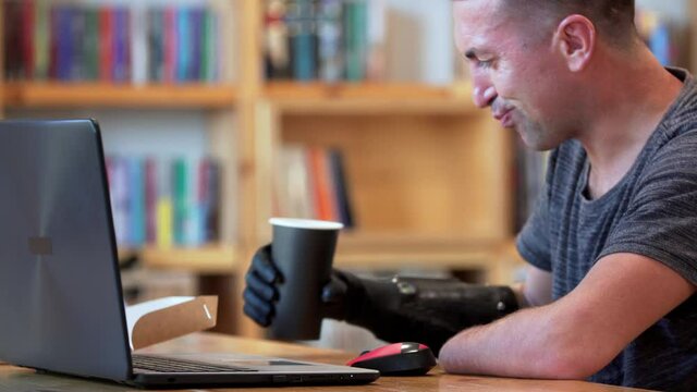 A man with amputated two stump hands is drinking coffee holding a cup with bionic hand and typing at the laptop. A man living with a disability learns to use the robotic prosthesis