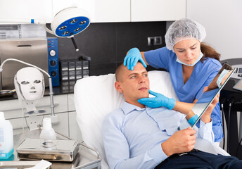 Skilled woman beautician consulting male client before facial treatment in medical esthetic office