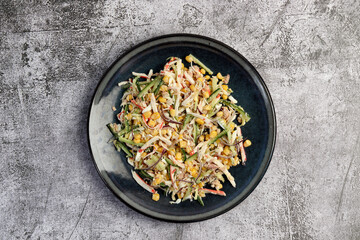 Tuna salad with surimi crab sticks, corn and cucumber on a round plate on a dark gray background....