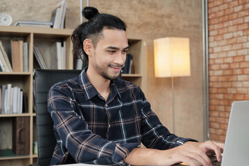 Fototapeta premium Young male worker of Asian ethnicity uses laptop to do creative work on white desk in front of bookshelf of a casual workplace, startup business person, and online e-commerce occupation.