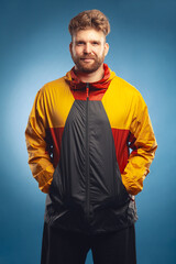 Outerwear for tourism and sports concept. Emotive portrait of laughing charismatic muscular 30-year-old man standing over blue background. Stylish haircut. Colourful jacket. Studio head shot