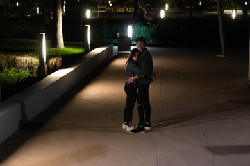 young latin couple having a romantic moment in the park at night, hugging, looking at each other, illuminated by the city lights.