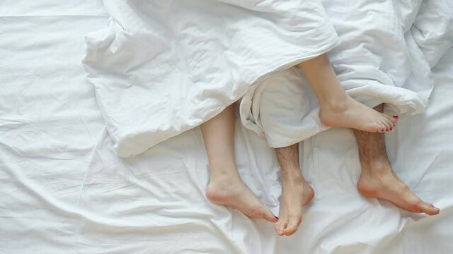 Young couple caresses each other after waking up on soft bed on white sheets sticking bare legs out of blanket in light bedroom at home, upper view.