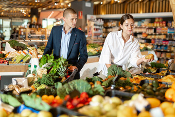 Positive young couple standing near the counter with a grocery cart in the supermarket, choose pears