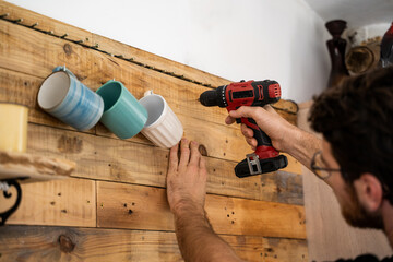 Man drilling hole to hang coffe mugs on wooden wall 