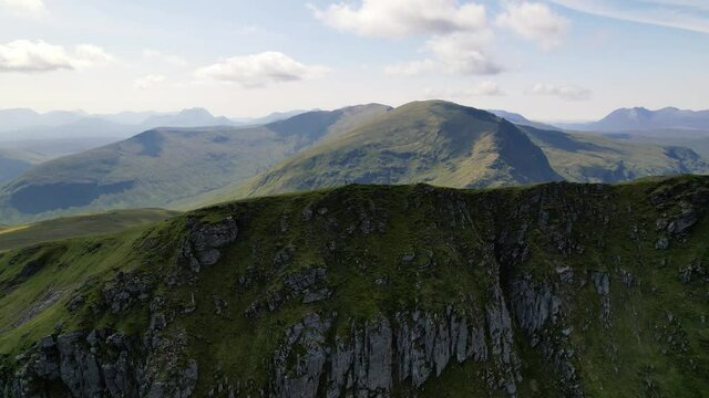 A Male standing on the top of green mountain ridge Fannichs between the peaks of the Scottish highlands on a sunny day. Slowly drone panning shot