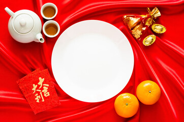 White plate on red satin cloth background with tea set, ingots, red bag (word means wealth), oranges and red envelope packets or ang bao(word means auspice) for Chinese new year concept.