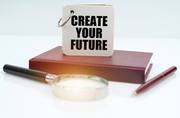 On a white surface lies a red notebook, a pen, a magnifying glass and a notebook with the inscription Create Your Future