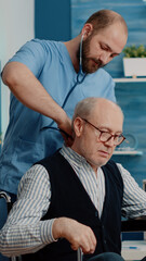 Fototapeta premium Nurse using stethoscope for consultation with disabled patient in nursing home. Medical assistant measuring heartbeat and doing healthcare checkup for retired man sitting in wheelchair.