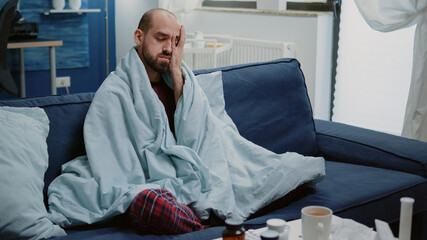 Unwell man having headache wrapped in blanket at home. Ill person with cold and flu symptoms...