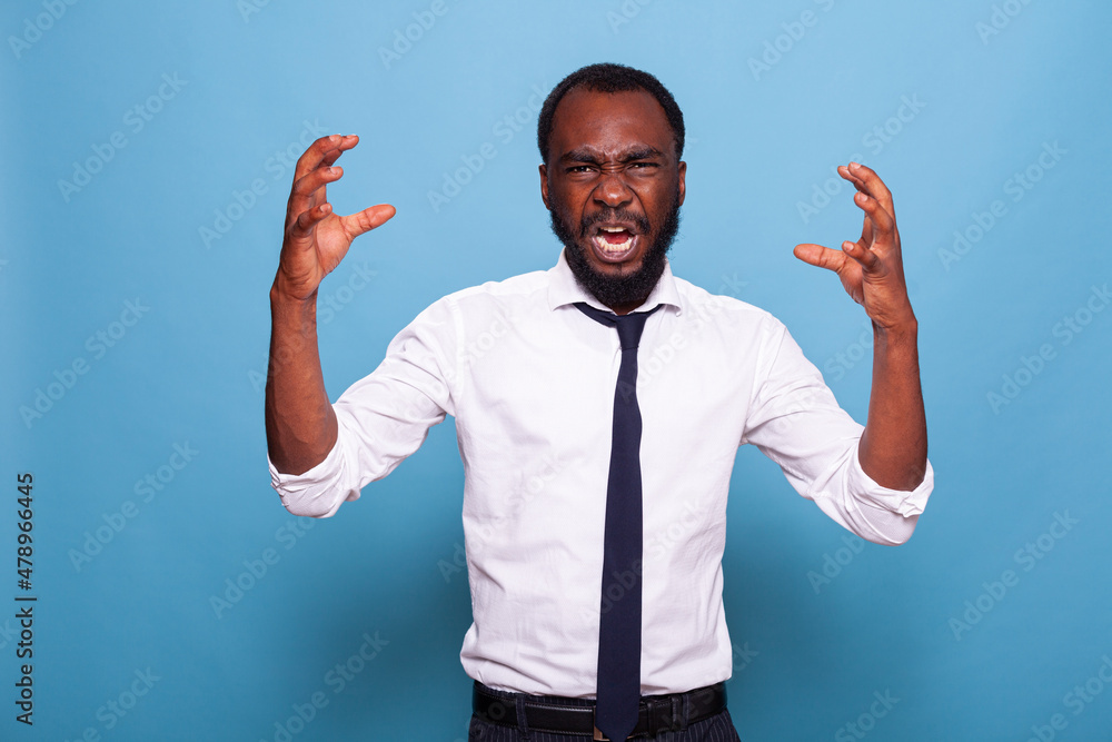 Wall mural portrait of upset angry african american businessman screaming and raising hands on blue background.