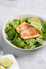 Grey bowl with fresh green raw spinach and fried red salmon, lime slices on grey concrete surface