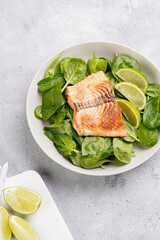 Grey bowl with fresh green raw spinach and fried red salmon, lime slices on grey concrete surface