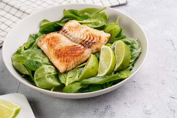 Grey bowl with fresh green raw spinach and fried red salmon, lime slices on grey concrete surface