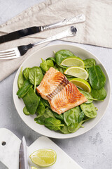 Grey bowl with fresh green raw spinach and fried red salmon, lime slices on grey concrete surface