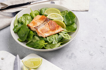 Grey bowl with fresh green raw spinach and fried red salmon, lime slices on grey concrete surface