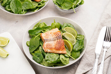 Grey bowl with fresh green raw spinach and fried red salmon, lime slices on grey concrete surface