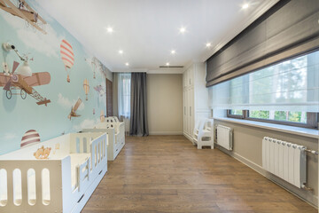 A spacious children's room with a blue wall decorated with drawings, a wooden floor, a large window and two white cots.