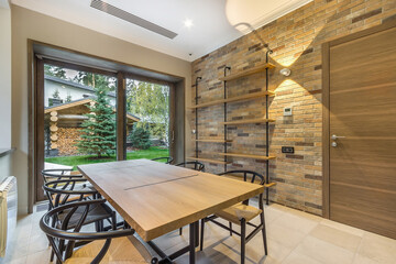 Stylish living room with brick wall and panoramic window in a new house. A large table with chairs in the center of the room.