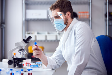 Male Lab Worker Wearing PPE Analysing Blood Samples In Laboratory With Microscope