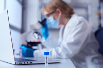 Female Lab Worker Wearing Lab Coat Working On Omicron Covid-19 Variant With Laptop And Microscope