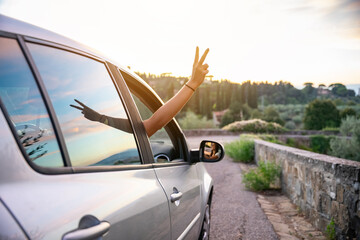 Two friends on holiday in the summer at sunset - Millennial show fingers up from the car window - Relaxed woman during a free trip in the nature - People happy for adventures and carefree leisure
