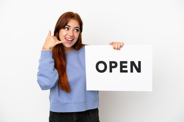 Young redhead woman isolated on white background holding a placard with text OPEN and doing phone gesture