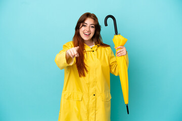 Redhead woman holding an umbrella isolated on blue background surprised and pointing front - obrazy, fototapety, plakaty