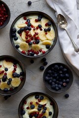 Breakfast bowl of yogurt, bananas, blue berries and pomegranate on a grey surface.  A spoon  and a napkin sits to the side of the bowls. 