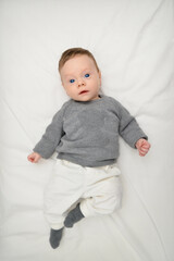 Baby boy smiling happy and looking curious at camera, lying on your back in the bed. Horizontal photo. Funny child infant with blue grey eyes.