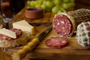 A sausage roll of wild boar salami sliced on a wooden board and being served with cheese and grapes on a wooden table.
