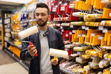 a man in a construction store picks up a roller for acrylic and oil paint next to a rack with tools for painting, a concept of home renovation and painting works