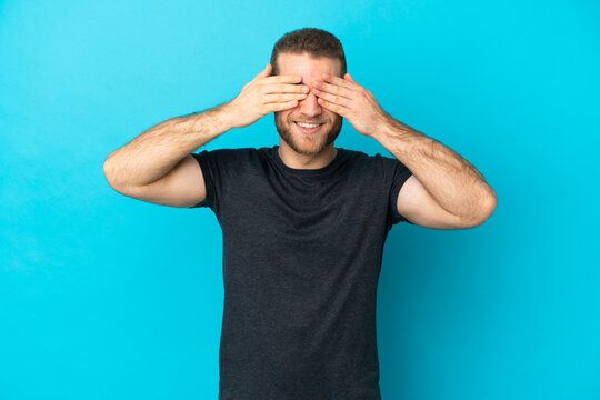 Young handsome caucasian man isolated on blue background covering eyes by hands and smiling