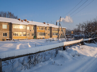 Fototapeta na wymiar Winter evening in a snowy village. The rays of the setting sun illuminate houses, fence, vegetable gardens and greenhouse.
