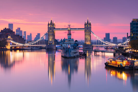 Sunrise view of HMS Belfast and Tower Bridge reflected in River Thames, with Canary Wharf in background, London