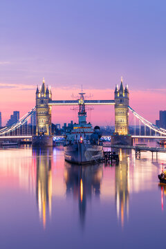 Sunrise view of HMS Belfast and Tower Bridge reflected in River Thames, with Canary Wharf in background, London