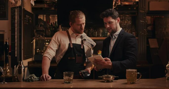 Caucasian male bartender standing behind bar speaking to manager holding digital tablet