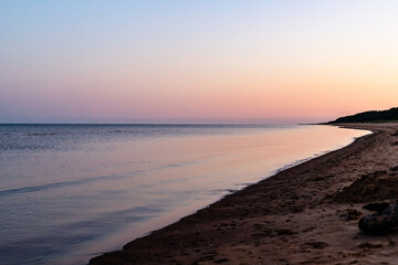 Fototapeta premium Sunrise on the beach during early morning. Calm smooth silky blue sea, light pink dawn, dark brown sand and dark forest at background. Nordic coastal daybreak at summer morning.