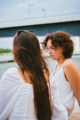 Two teenage girl talking and spending a relax time. They are sitting by a river and there is a blue bridge at the background.