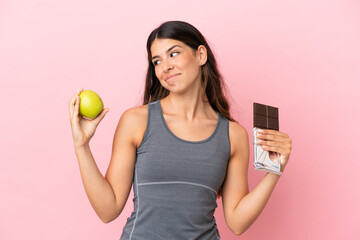 Young caucasian woman isolated on pink background having doubts while taking a chocolate tablet in...
