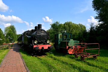 Fototapeta na wymiar A view of a well maintained public park full of shrubs, trees, and grass, with a train station located nearby and an old traditional steam locomotive waiting for departure seen on a sunny summer day
