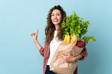 Young woman holding a grocery shopping bag isolated on blue background pointing back