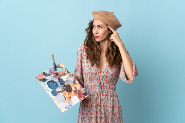 Young artist girl holding a palette isolated on blue background having doubts and thinking © luismolinero