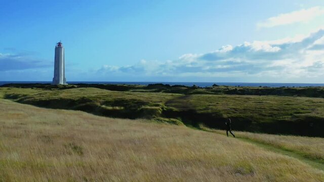 Girl walking the grass path to the lighthouse in the field on a cliff of Iceland