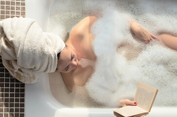 Young woman takes a bath with lush foam near the window reading the book. Personal hygiene,...