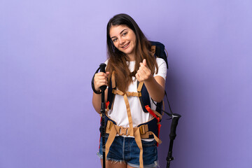 Young woman with backpack and trekking poles isolated on purple background making money gesture