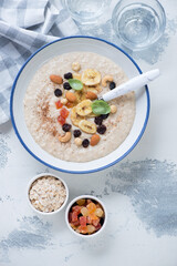 Plate of oatmeal porridge with dried banana, candied fruits and nuts, flatlay on a white concrete background, vertical shot