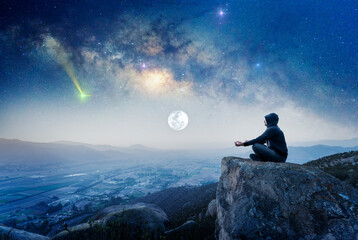 person on the mountain outdoors meditating or praying at night with Milky Way and Moon background.