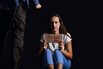 Aggressive man and scared young woman holding paper with text PROTECT ME on dark background. Violence concept