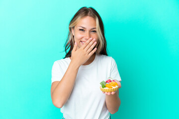 Young Russian woman holding a fruit sweet isolated on blue background happy and smiling covering mouth with hand